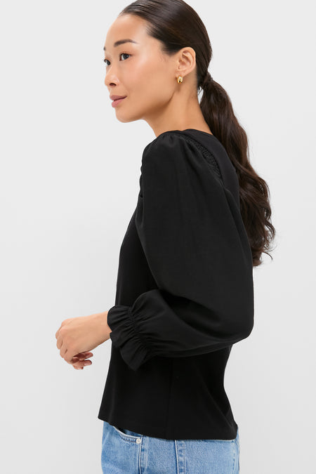 A woman with long dark hair in a ponytail models a Pomander Place black puff sleeves blouse and light blue jeans against a plain light background.