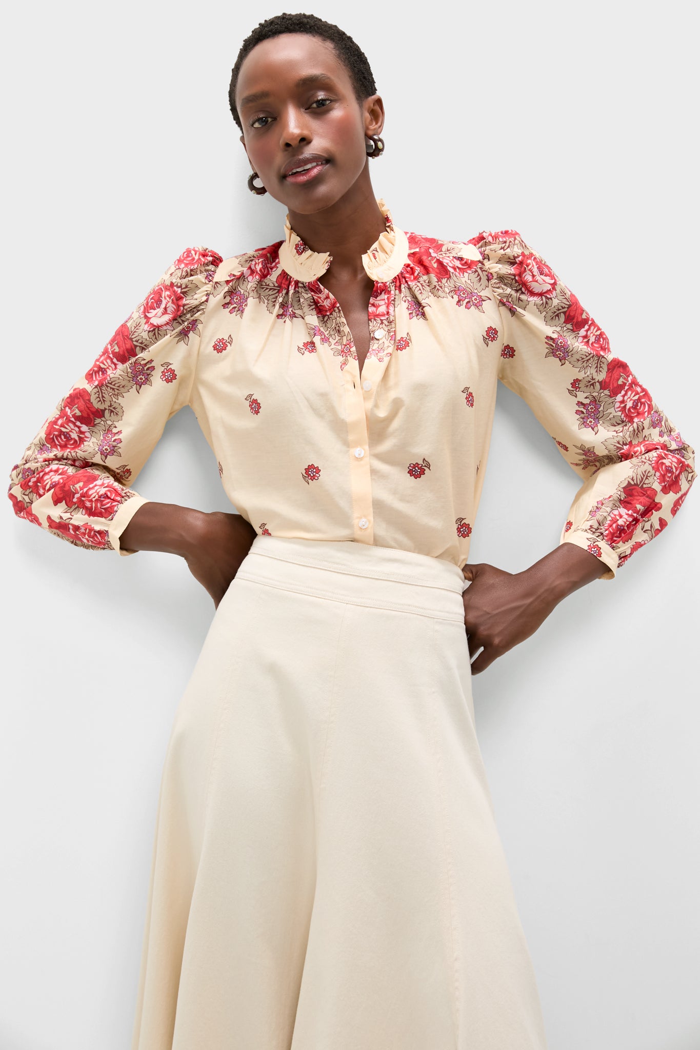 A person stands against a plain light background wearing an Alix of Bohemia boho-chic blouse with red floral embroidery on the shoulders and sleeves, paired with a cream skirt.