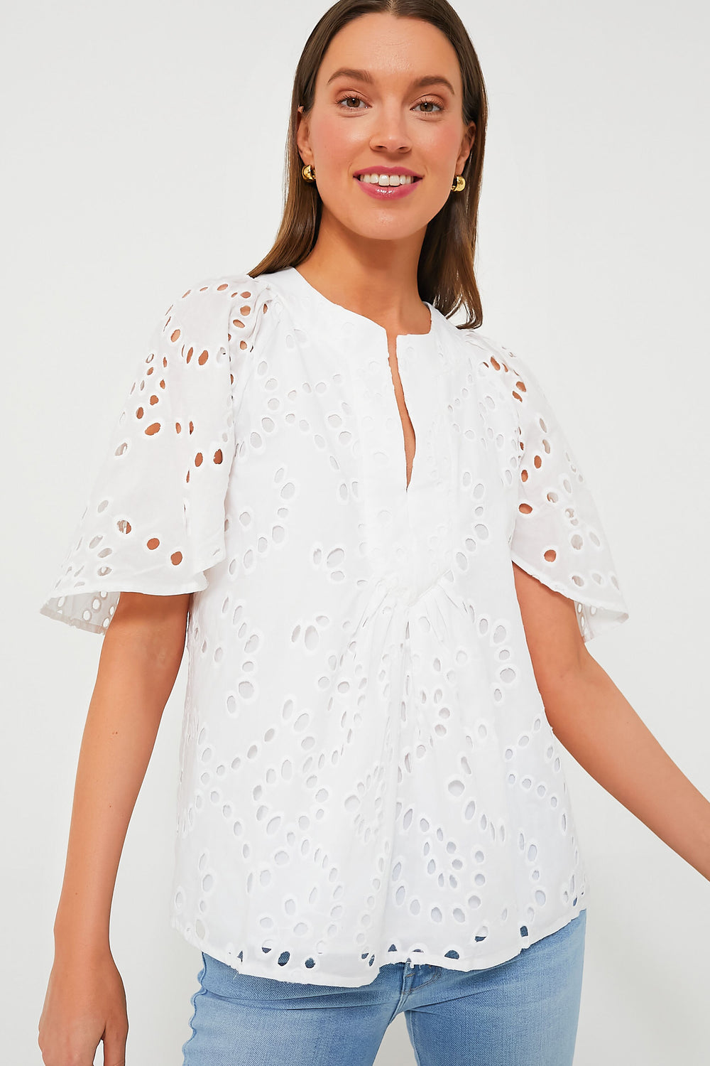 A woman smiles brightly in a Tuckernuck white eyelet short-sleeve blouse with flutter sleeves and light blue jeans.