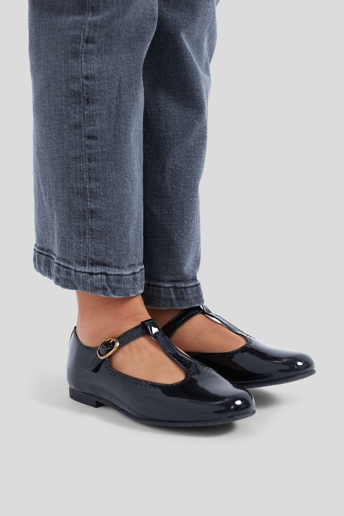 A child in dark blue jeans and La Coqueta black patent leather T-Bar shoes stands on a light grey background.