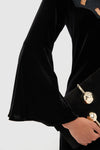 Close-up of Accademia Black Velvet Bell Sleeve Catherine Maxi Dress, featuring lace yoke and flared sleeves, paired with a high-end black suede clutch with gold accents.