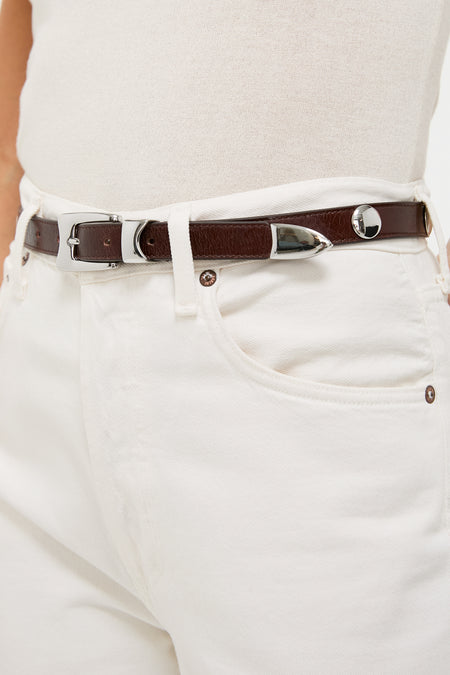 Close-up of Sepia Aiden Studded Rugous Belt in brown leather, featuring a polished silver buckle and studs, paired with cream jeans for a classic, effortless look.