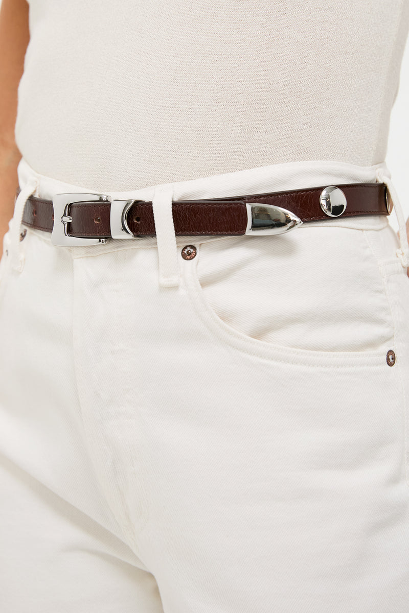 Close-up of Sepia Aiden Studded Rugous Belt in brown leather, featuring a polished silver buckle and studs, paired with cream jeans for a classic, effortless look.