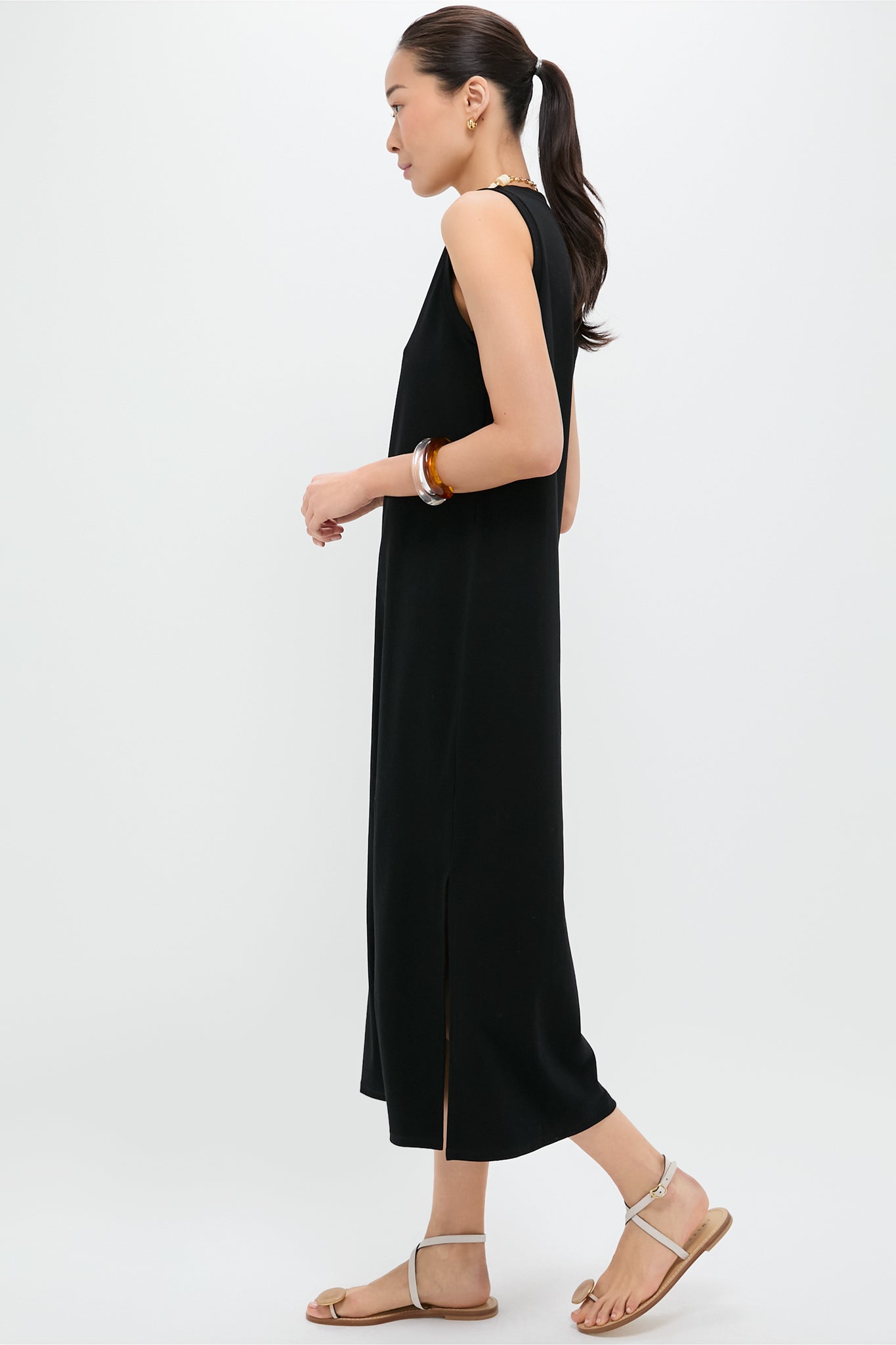 A woman in a Pomander Place sleeveless black cotton-spandex maxi dress with side slits, sandals, and bracelets stands in profile against a plain light background.