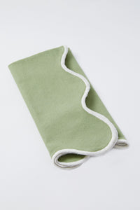 A folded light green Tuckernuck Home blanket with white trim, displayed on a white surface, pairs beautifully with scalloped napkins for an elegant touch.