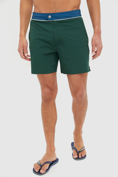 A person wearing Spruce dark green mens surf shorts featuring a blue waistband with button, paired with blue flip-flops.