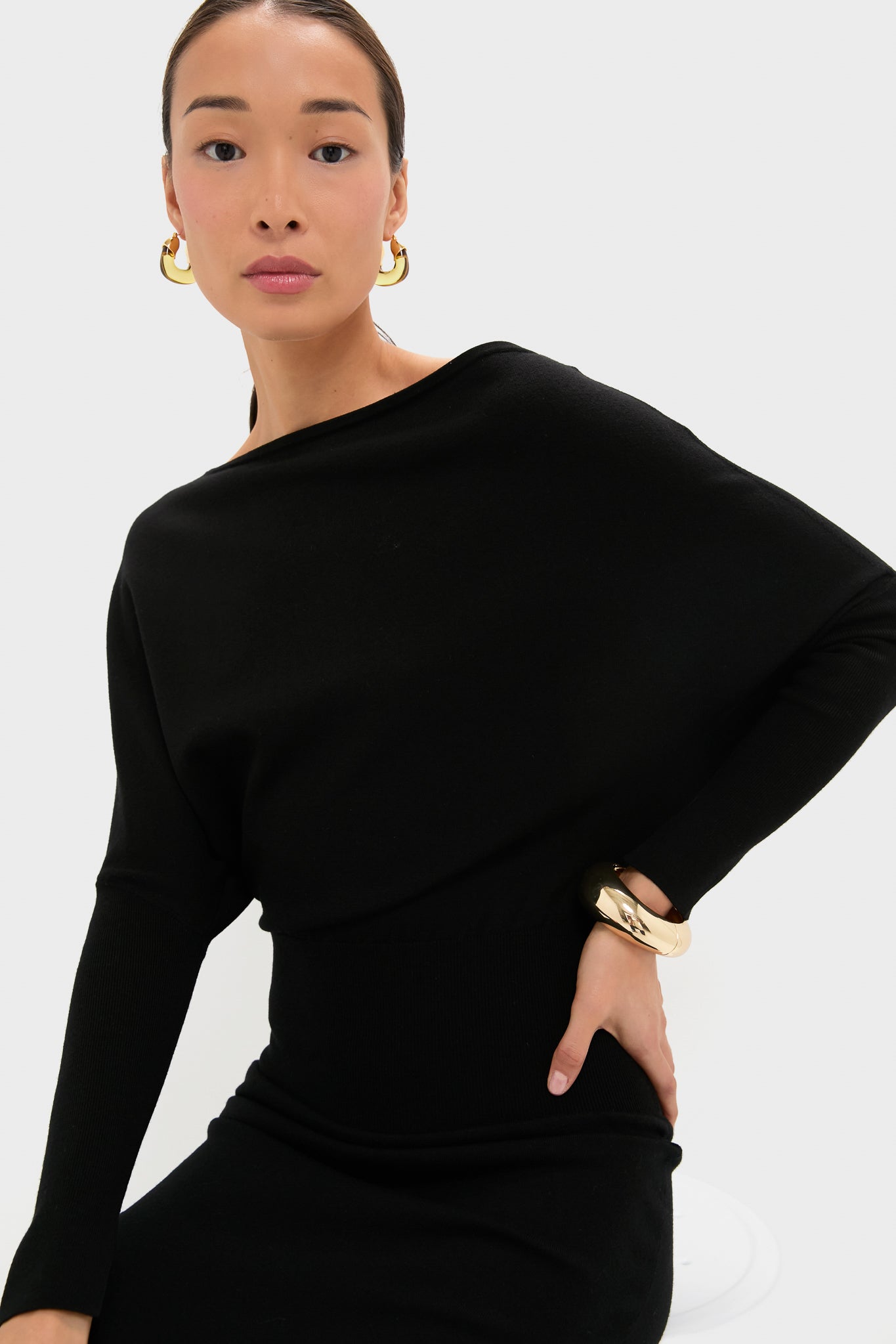 A woman wearing a black SIMKHAI luxury long-sleeve dress, gold hoop earrings, and a gold bracelet sits on a stool against a plain white background.