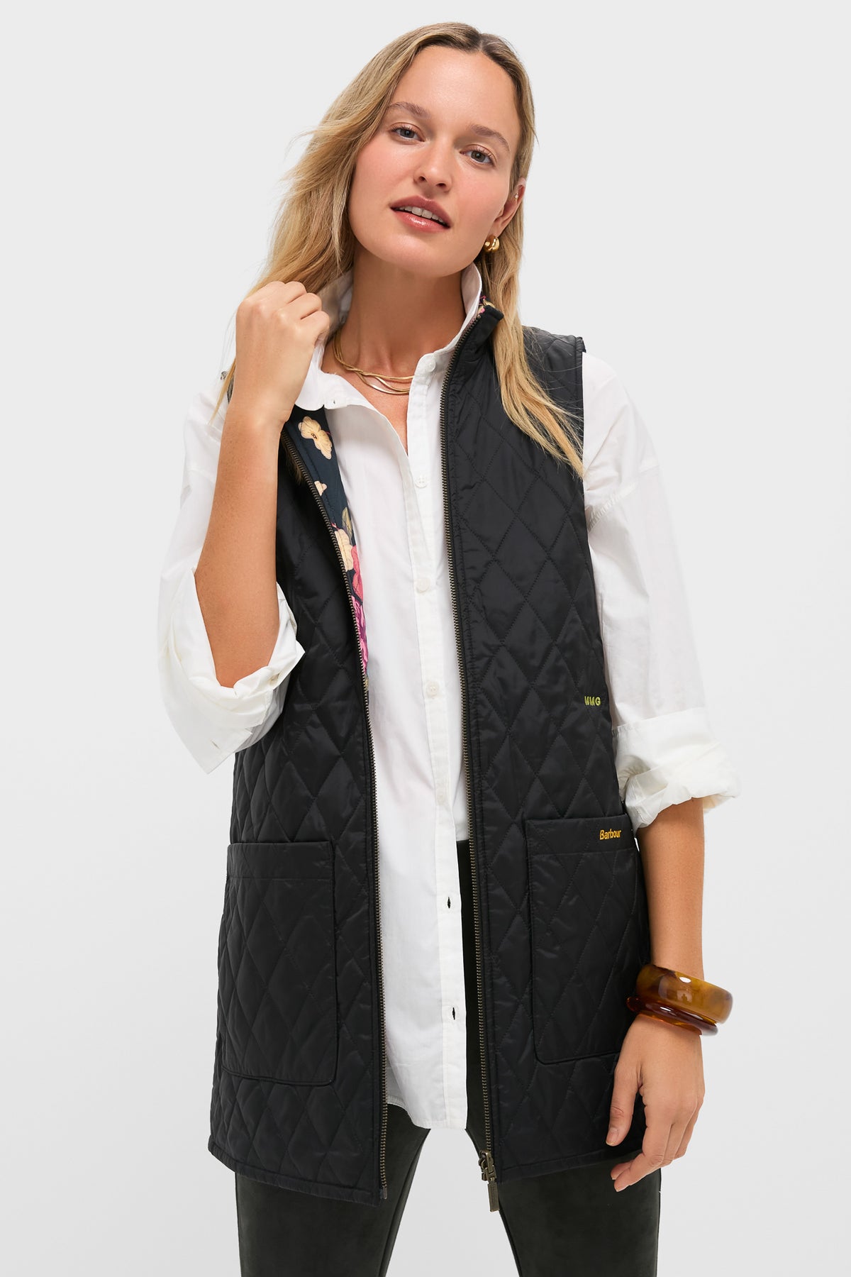 A woman wears a white button-up shirt and a Barbour x Tuckernuck black quilted vest with front pockets and a reversible Paisley liner, posing against a plain light background.