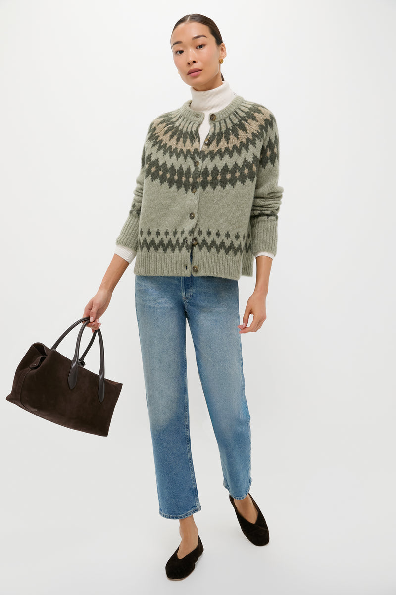 A person wearing a patterned green Fair Isle cardigan by Tuckernuck, a white turtleneck, blue jeans, black flats, and holding a black handbag stands against a plain white background.
