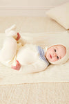 A baby rests on a cream blanket, wearing a cozy Pepa London Fair Isle baby set made from soft merino wool with blue details and a matching hat, on a light carpeted floor.