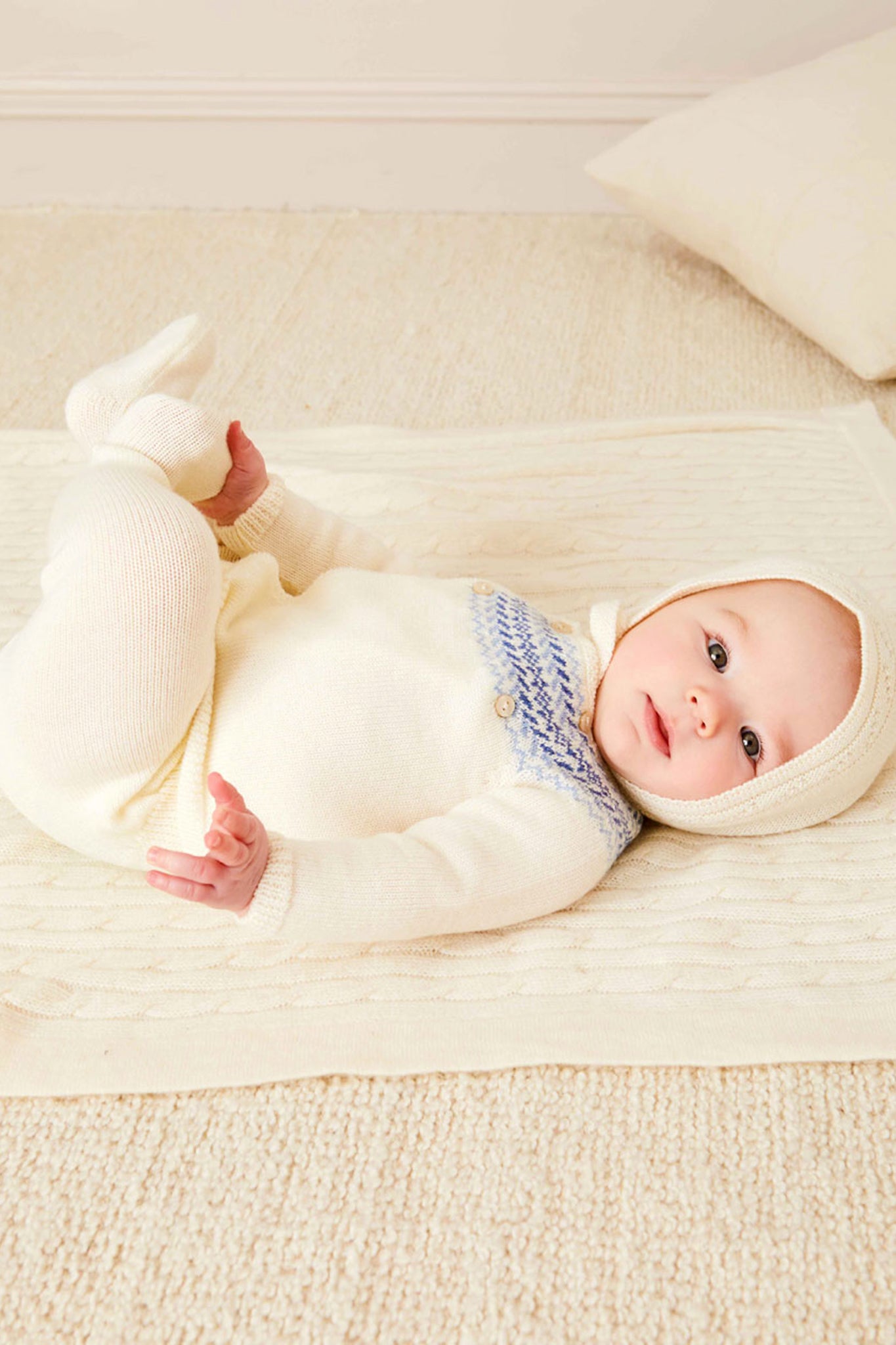 A baby rests on a cream blanket, wearing a cozy Pepa London Fair Isle baby set made from soft merino wool with blue details and a matching hat, on a light carpeted floor.