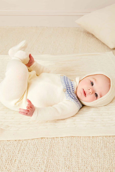 A baby rests on a cream blanket, wearing a cozy Pepa London Fair Isle baby set made from soft merino wool with blue details and a matching hat, on a light carpeted floor.