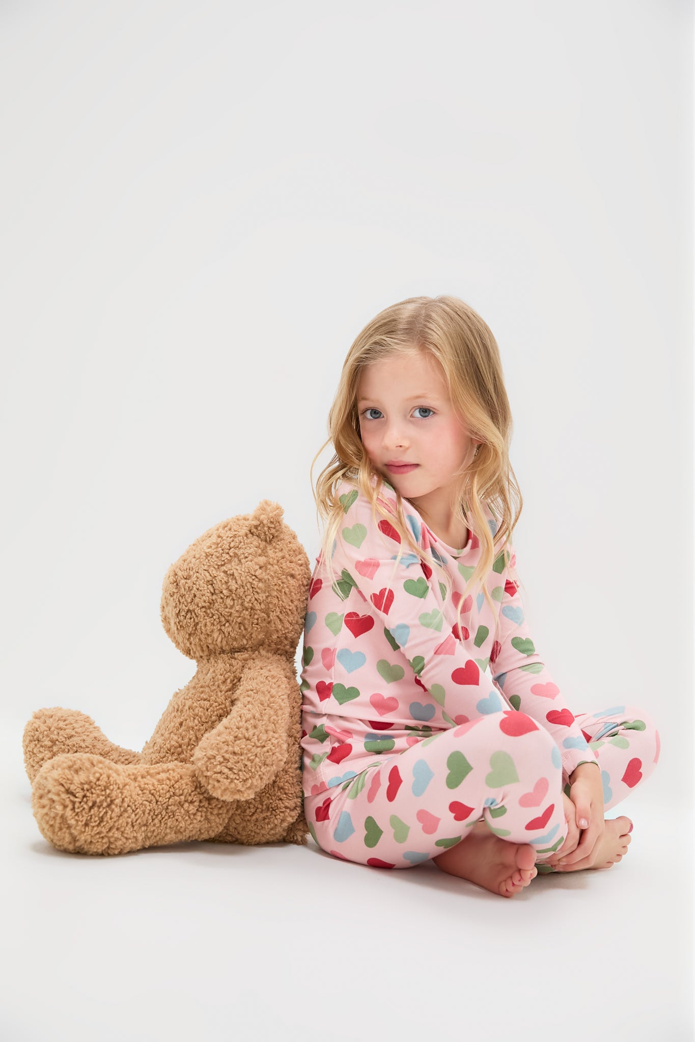 Young girl in Pink Chicken Small Hearts Kids Bamboo Pj Set, lounging against a teddy bear. The classic, effortless pajamas feature a fun, heart-print design, perfect for high-end comfort.