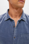 A close-up of a person wearing a blue Marine Layer cotton corduroy button-down shirt with a collar and chest pocket; their face is partially out of frame.