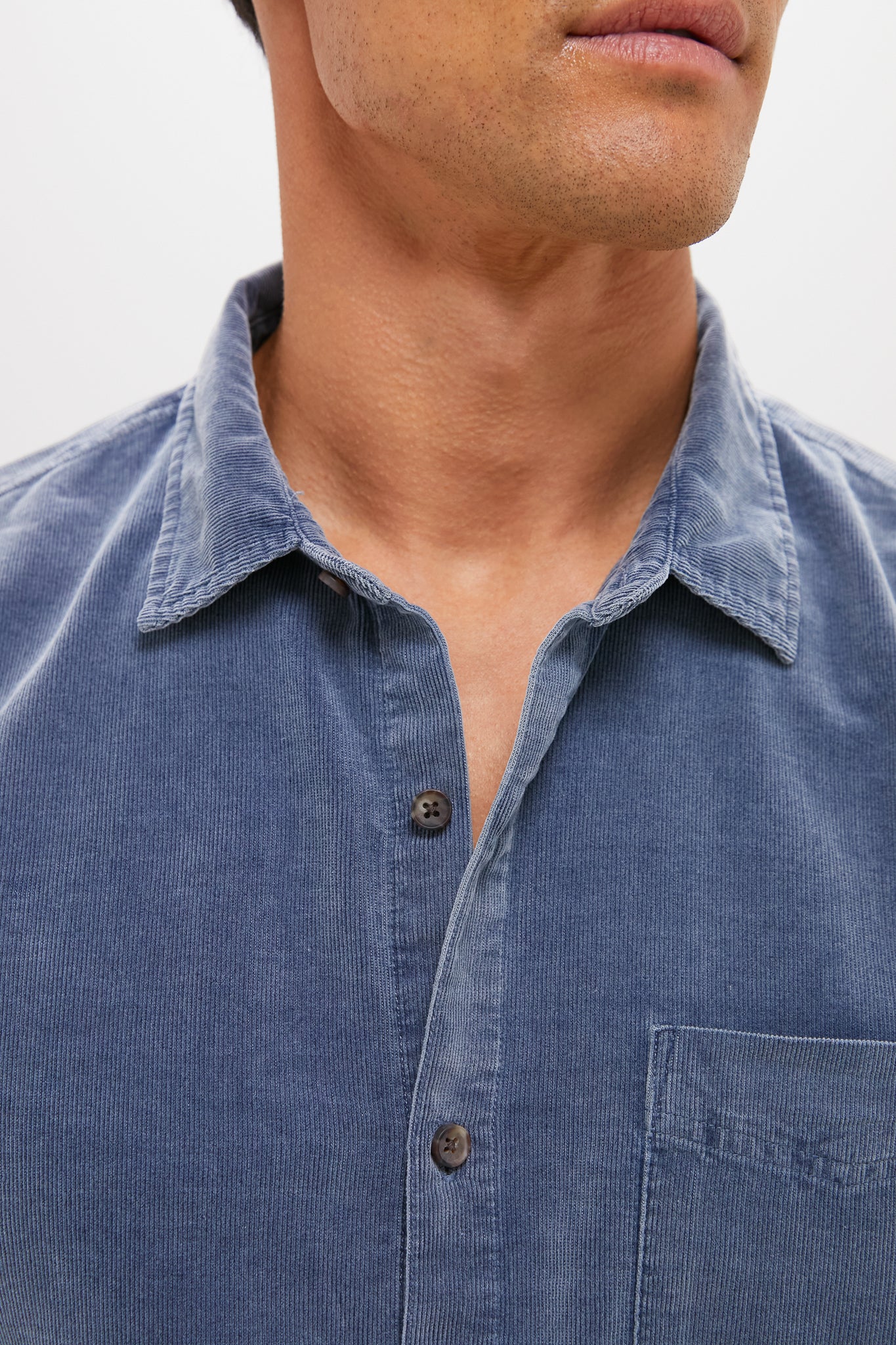 A close-up of a person wearing a blue Marine Layer cotton corduroy button-down shirt with a collar and chest pocket; their face is partially out of frame.