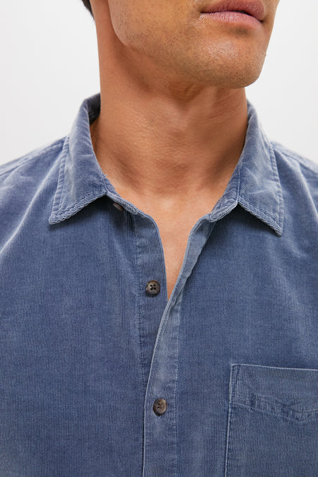 A close-up of a person wearing a blue Marine Layer cotton corduroy button-down shirt with a collar and chest pocket; their face is partially out of frame.