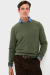 A man stands confidently in a green 100% wool Barbour sweater over a blue collared shirt, paired with brown corduroy pants and a brown belt.