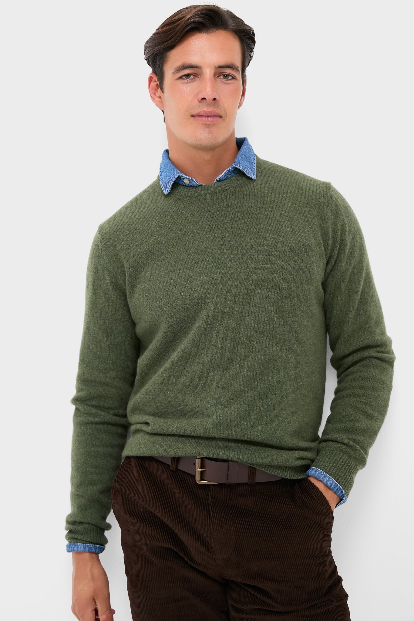 A man stands confidently in a green 100% wool Barbour sweater over a blue collared shirt, paired with brown corduroy pants and a brown belt.