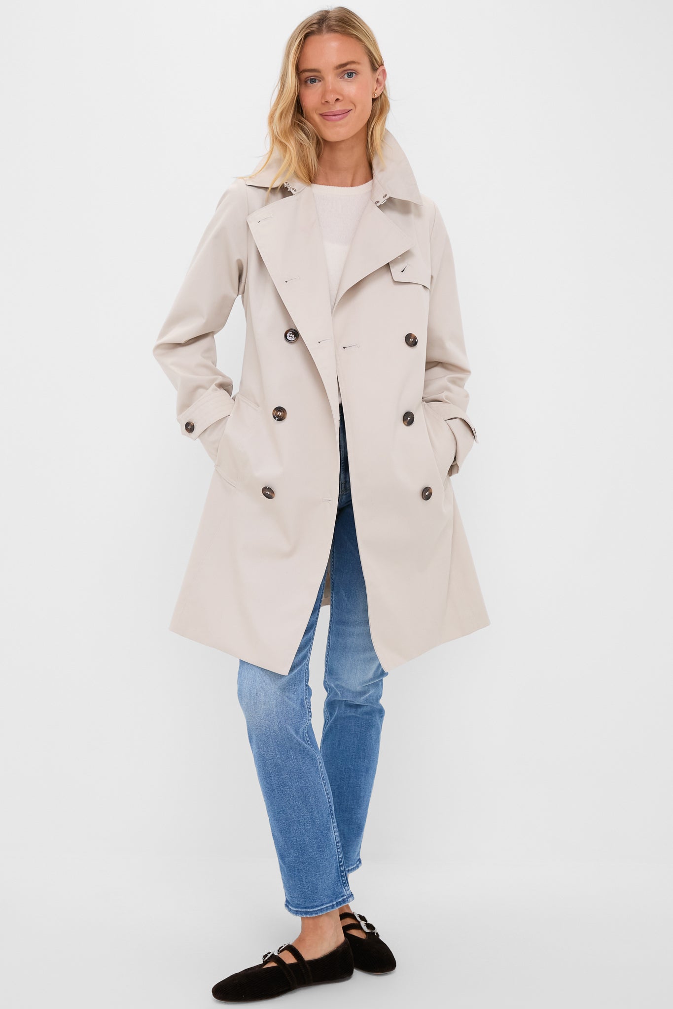 Light-skinned woman wearing the Mist Short Greta Showerproof Jacket, a classic double-breasted beige trench from J. Barbour® & Sons, paired with jeans and Mary Jane shoes.