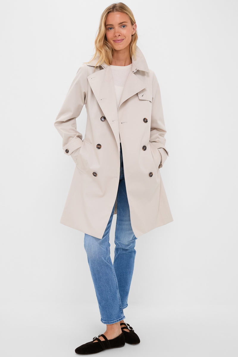 Light-skinned woman wearing the Mist Short Greta Showerproof Jacket, a classic double-breasted beige trench from J. Barbour® & Sons, paired with jeans and Mary Jane shoes.