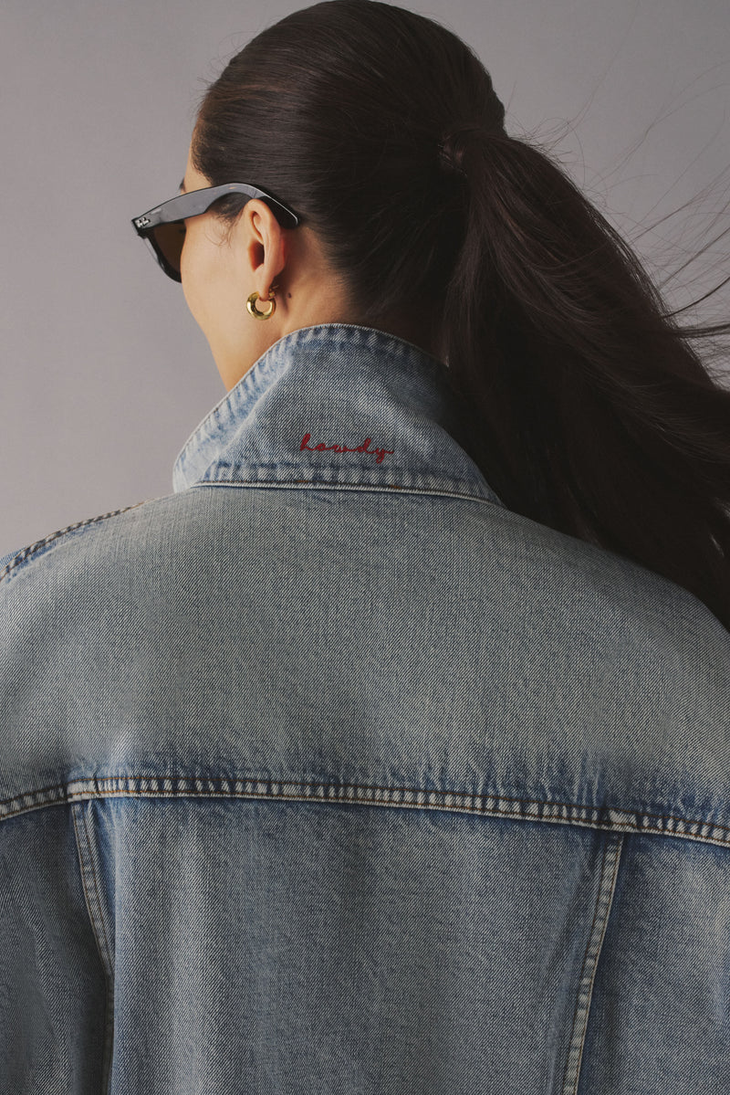 Back of a woman with a ponytail wearing a denim jacket with the collar popped. On the collar there's red script that says "howdy." 