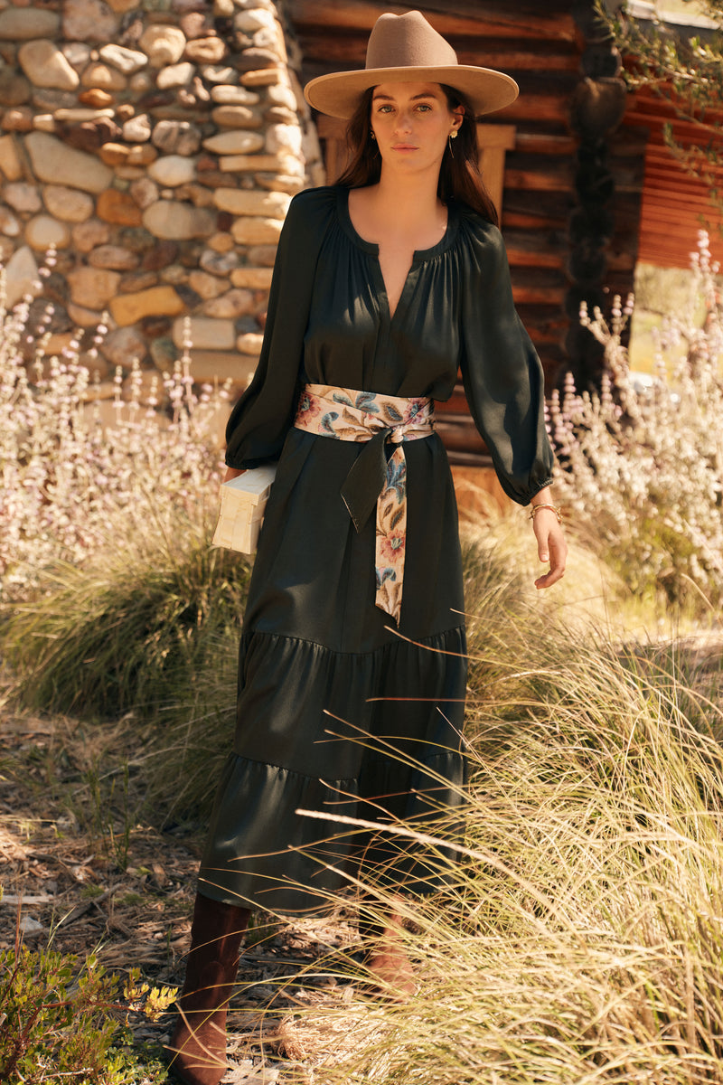 Woman outside with a stone and wood cabin in the background. She's wearing a tan cowboy hat, a long sleeved green dress with a contrasting floral belt, and a gold bracelet and earrings. She's holding a pearlescent clutch. 