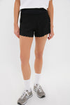 A person wears Tnuck Sport black scalloped hem athletic shorts with compression lining, white socks, gray sneakers, and a white top.