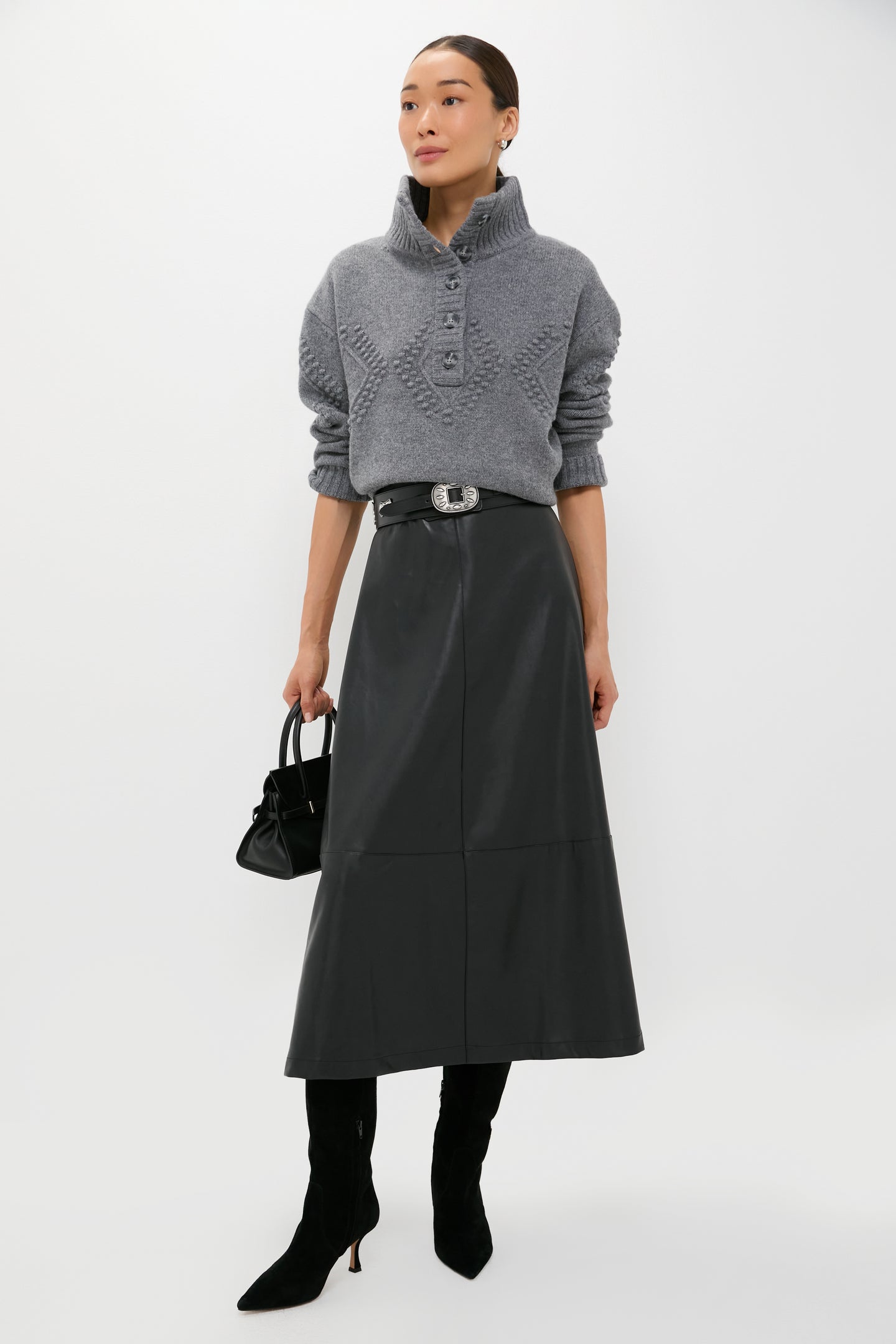A woman wears a Tuckernuck gray knit sweater, black leather belted skirt, black heeled boots, and carries a small black handbag against a white backdrop—chic office style.