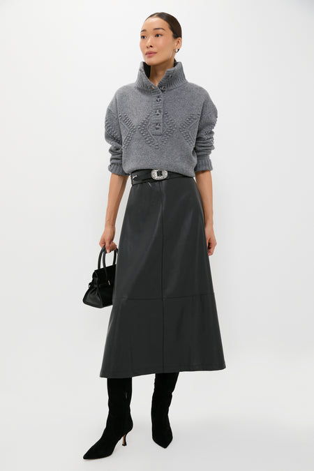 A woman wears a Tuckernuck gray knit sweater, black leather belted skirt, black heeled boots, and carries a small black handbag against a white backdrop—chic office style.