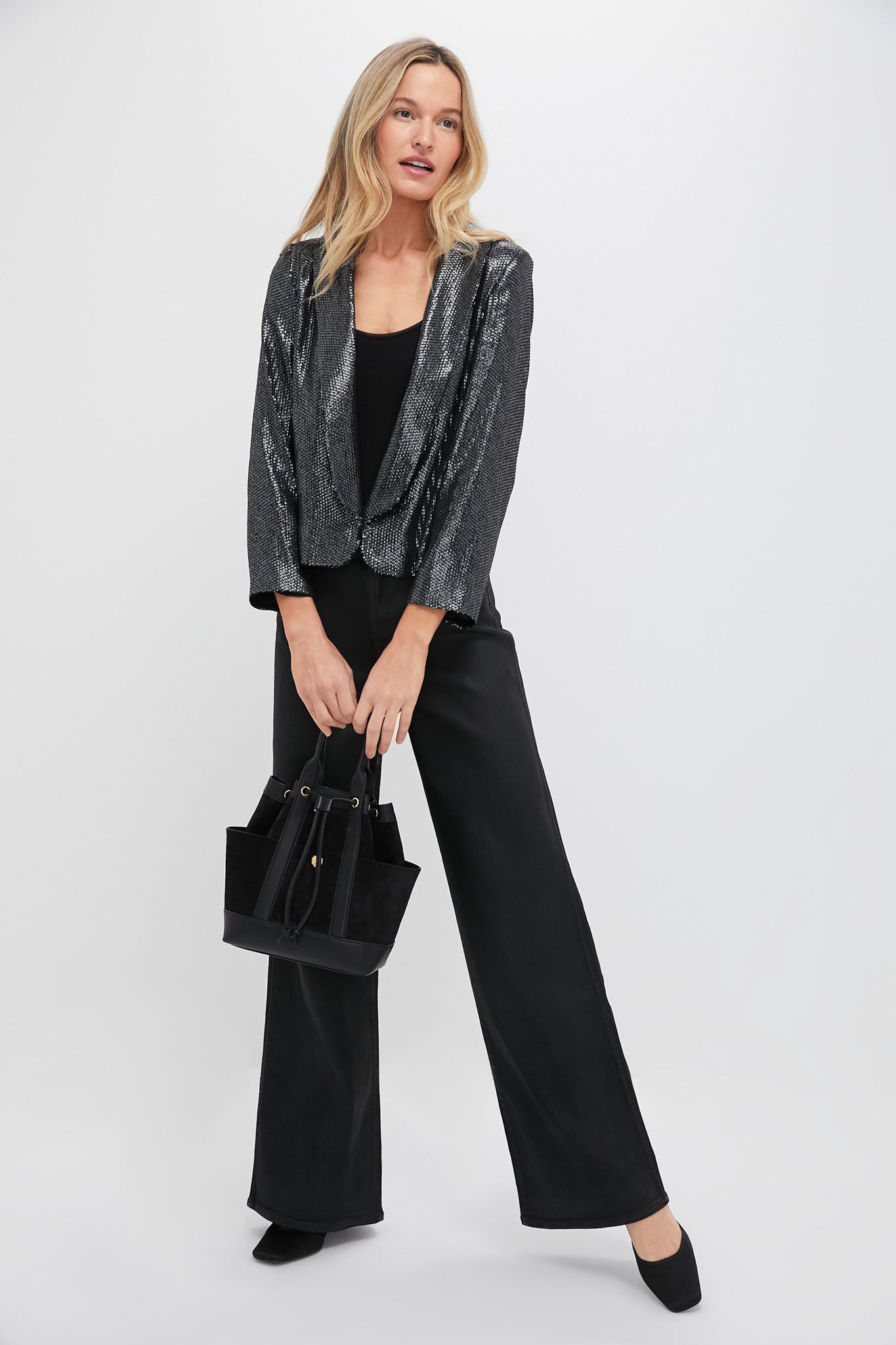 A woman stands against a plain background wearing a Tuckernuck gunmetal sequin jacket, black top, black wide-leg pants, black heels, and holding a black handbag.