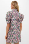 A woman faces away, wearing a Hyacinth House floral mini dress with puffed short sleeves and a ruffled collar—an ideal resort choice in pink, green, and purple blooms.