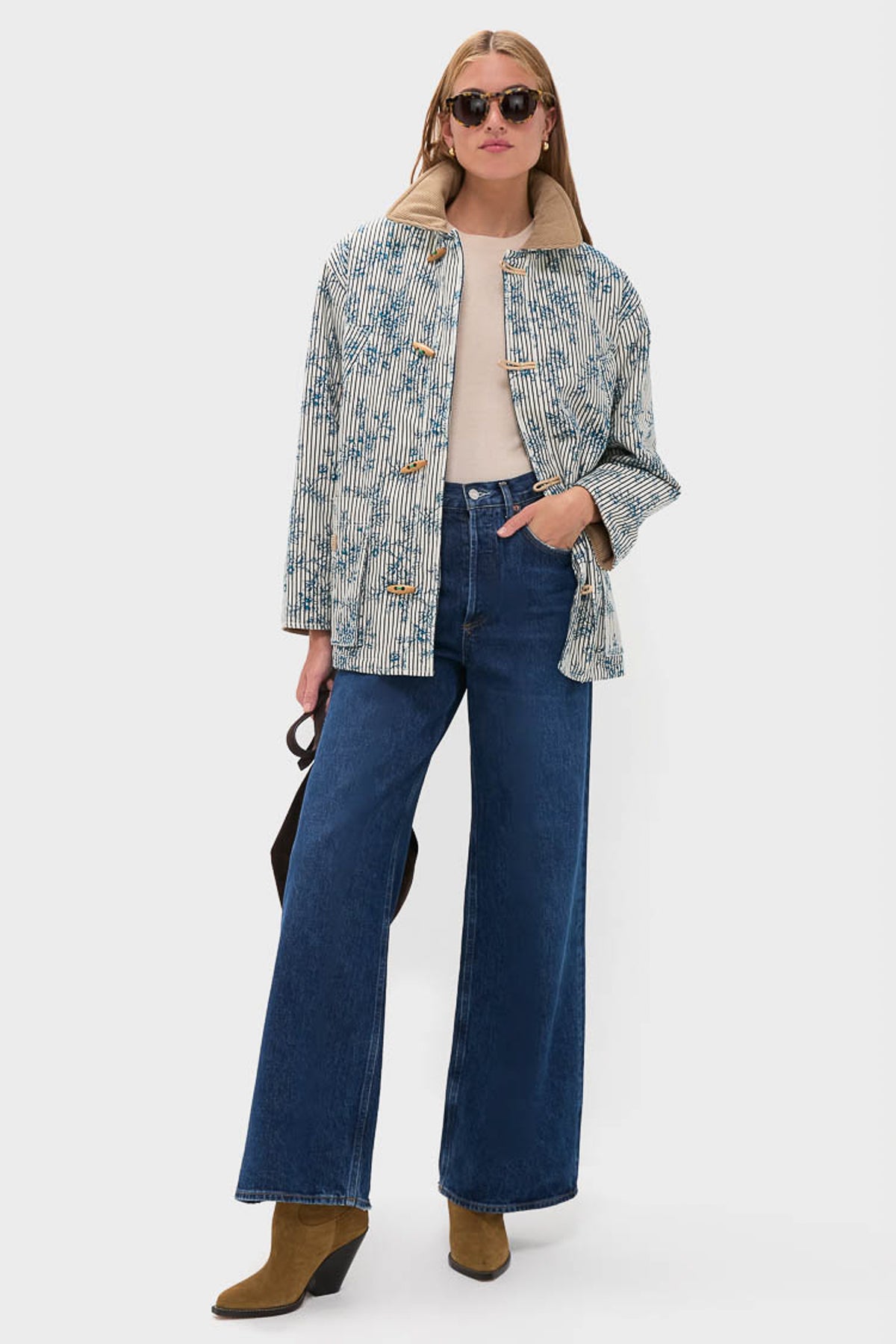 A person wearing Hunter Bell sunglasses, an oversized barn jacket, beige top, blue Italian stretch cotton denim wide-leg jeans, and brown heeled boots stands holding a dark-colored handbag.