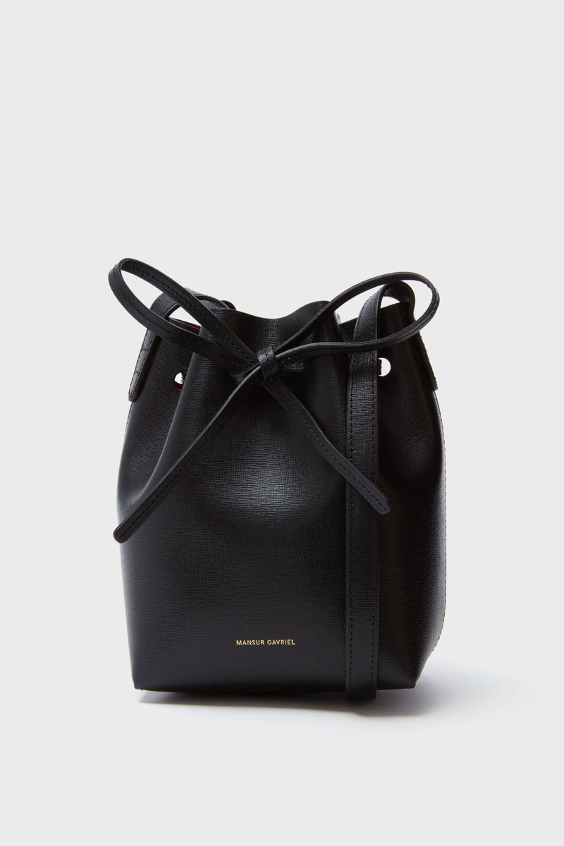 A black Mansur Gavriel Mini Mini Bucket Bag made from Italian Saffiano leather, featuring a drawstring closure and gold-embossed branding near the bottom.
