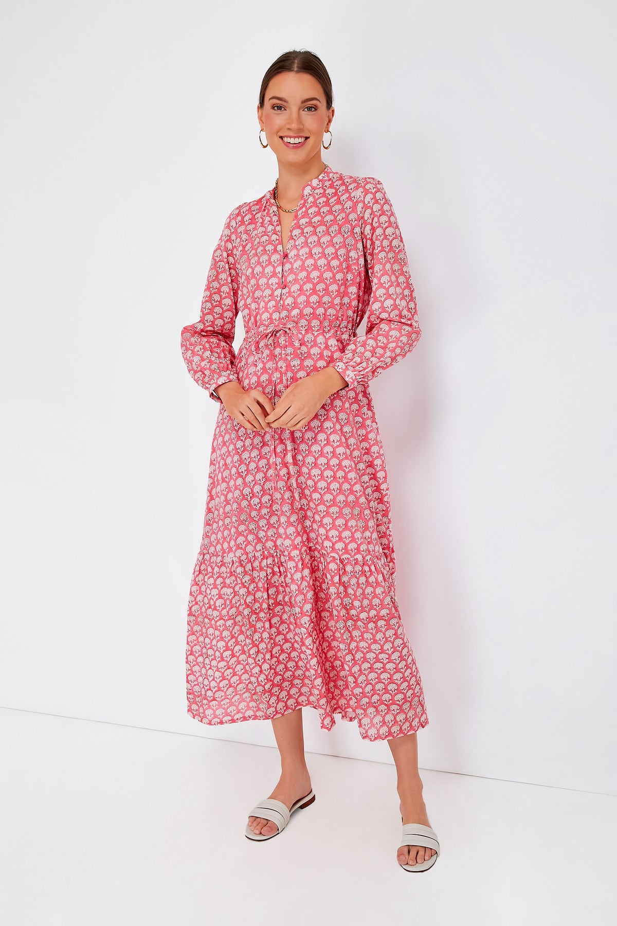 A woman wears a long-sleeved, pink patterned hand-block printed maxi dress by Love Tomas by Emerson Fry paired with white slide sandals for a boho-chic look.