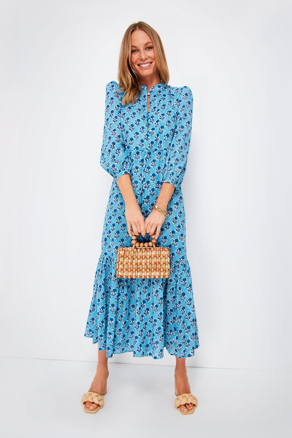 Woman in a blue floral boho dress from Love Tomas by Emerson Fry, carrying a small woven handbag and wearing beige sandals.