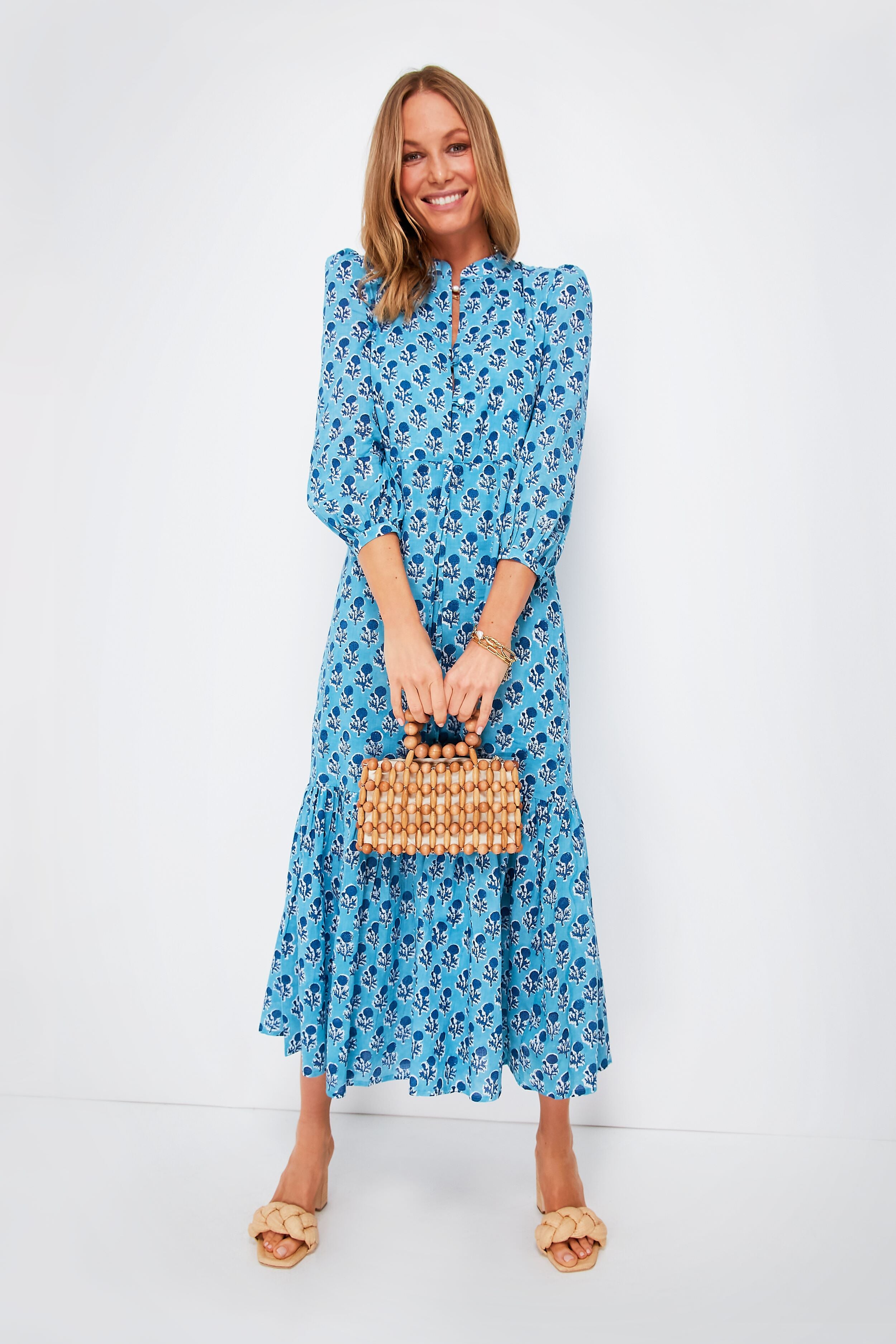 Woman in a blue floral boho dress from Love Tomas by Emerson Fry, carrying a small woven handbag and wearing beige sandals.
