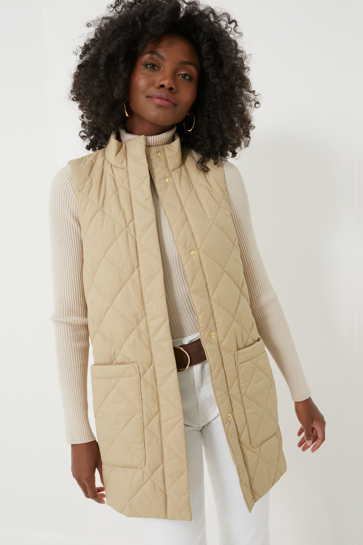 A woman wears a beige Barbour diamond-quilted Cosmia Liner vest over a light ribbed turtleneck, styled with white pants and a brown belt.
