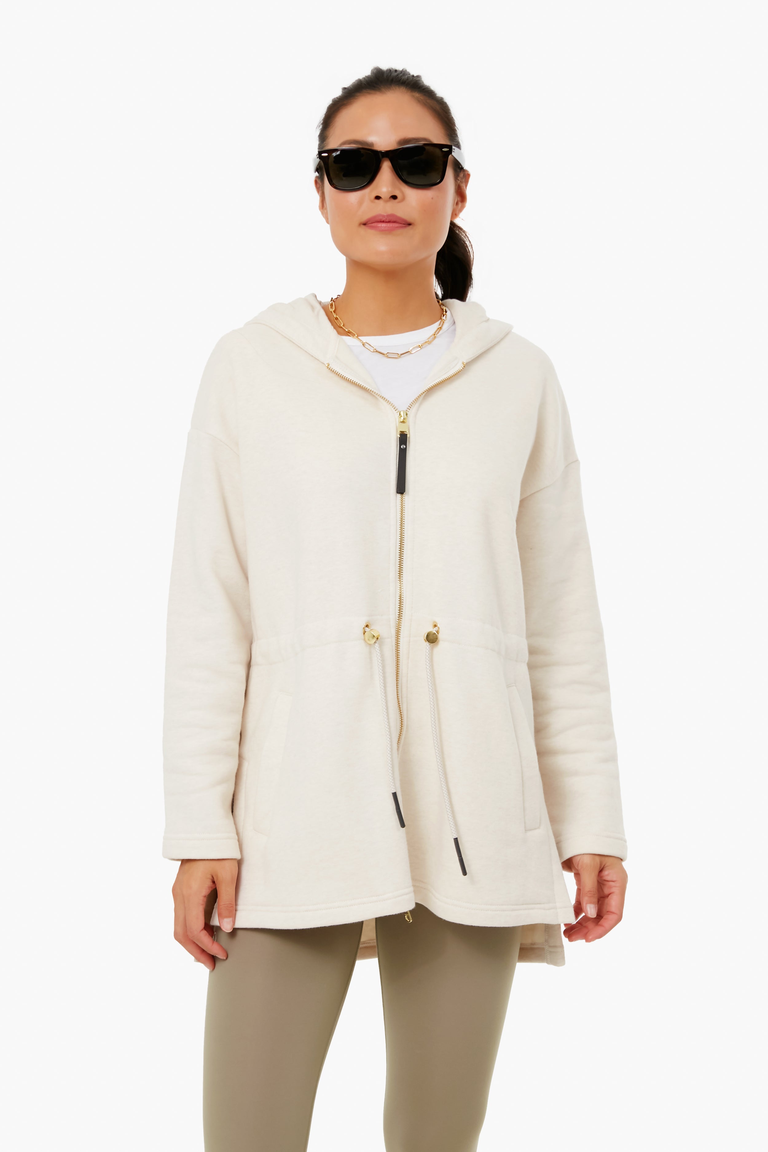 A woman wearing sunglasses, a cream oversized longline Varley Victoria Hoodie with drawstring waist, and light olive leggings showcases a performance-fashion look.