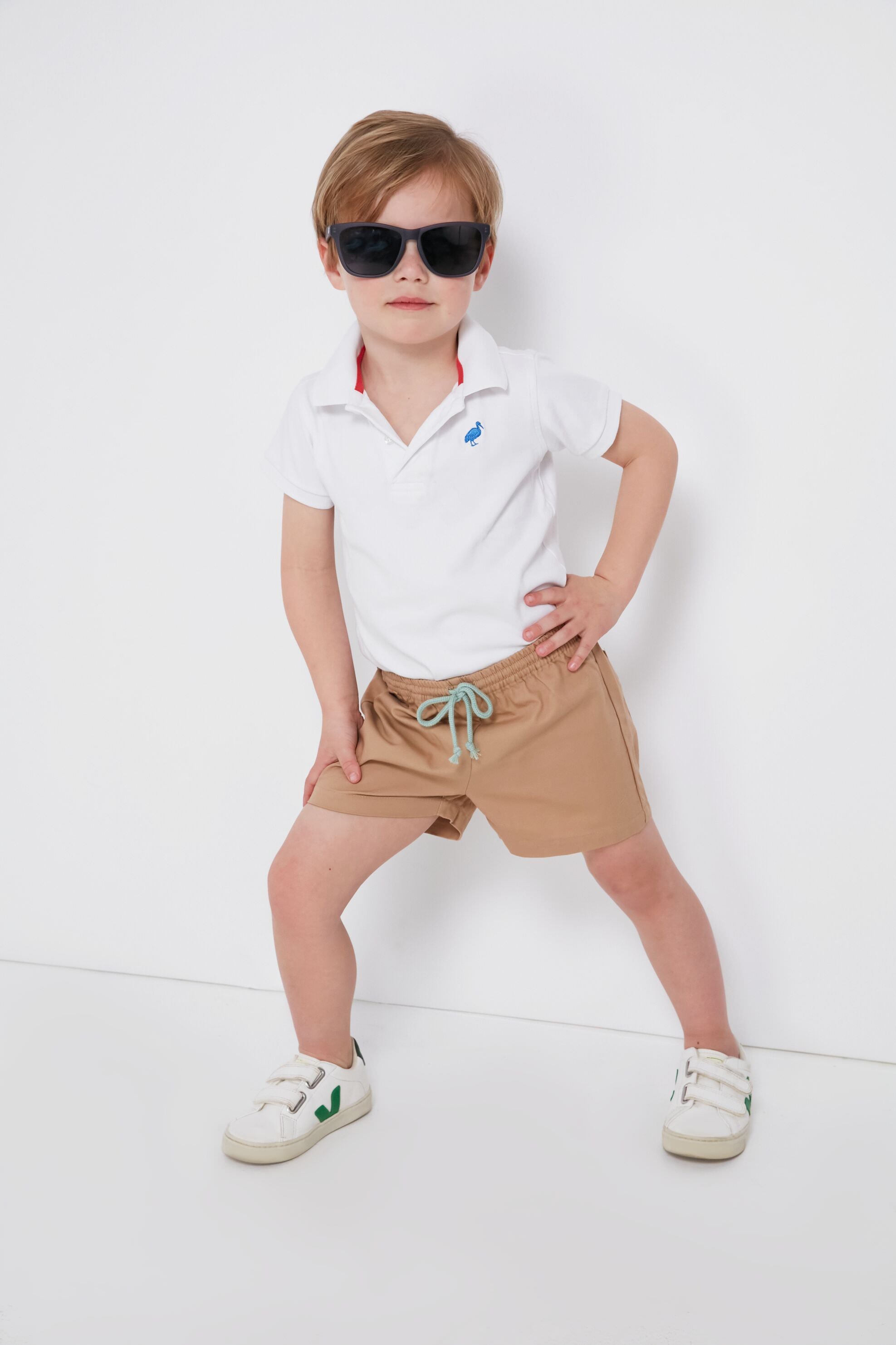 A young child stands confidently with one hand on hip, wearing Oso & Me sunglasses, a white polo shirt, adjustable waist shorts, and white sneakers—showcasing stylish and durable children's clothing.