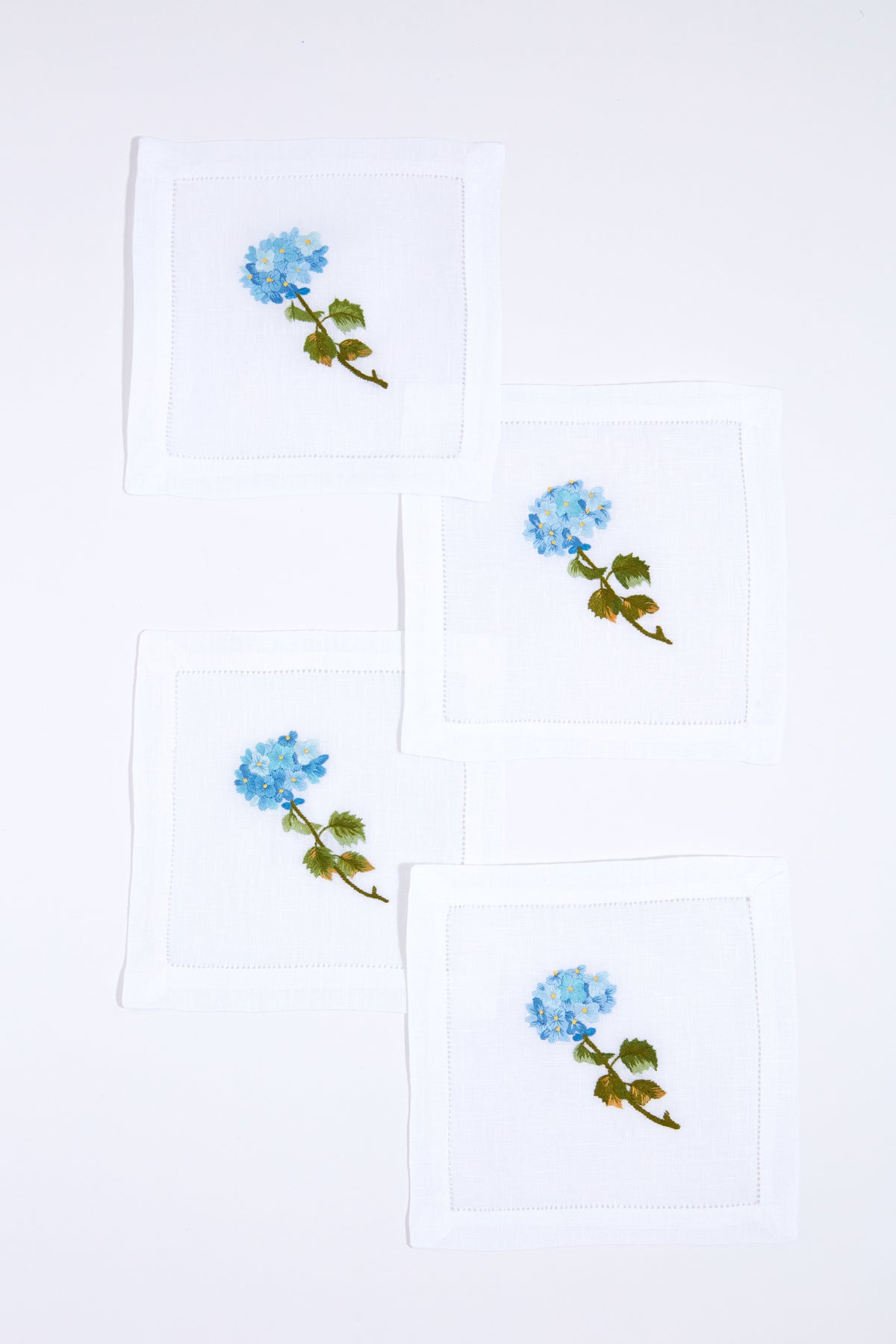 Set of four white linen napkins by Tuckernuck Home, each hand-embroidered with a blue flower and green leaves. Ideal as hydrangea-themed cocktail napkins or a thoughtful hostess gift.