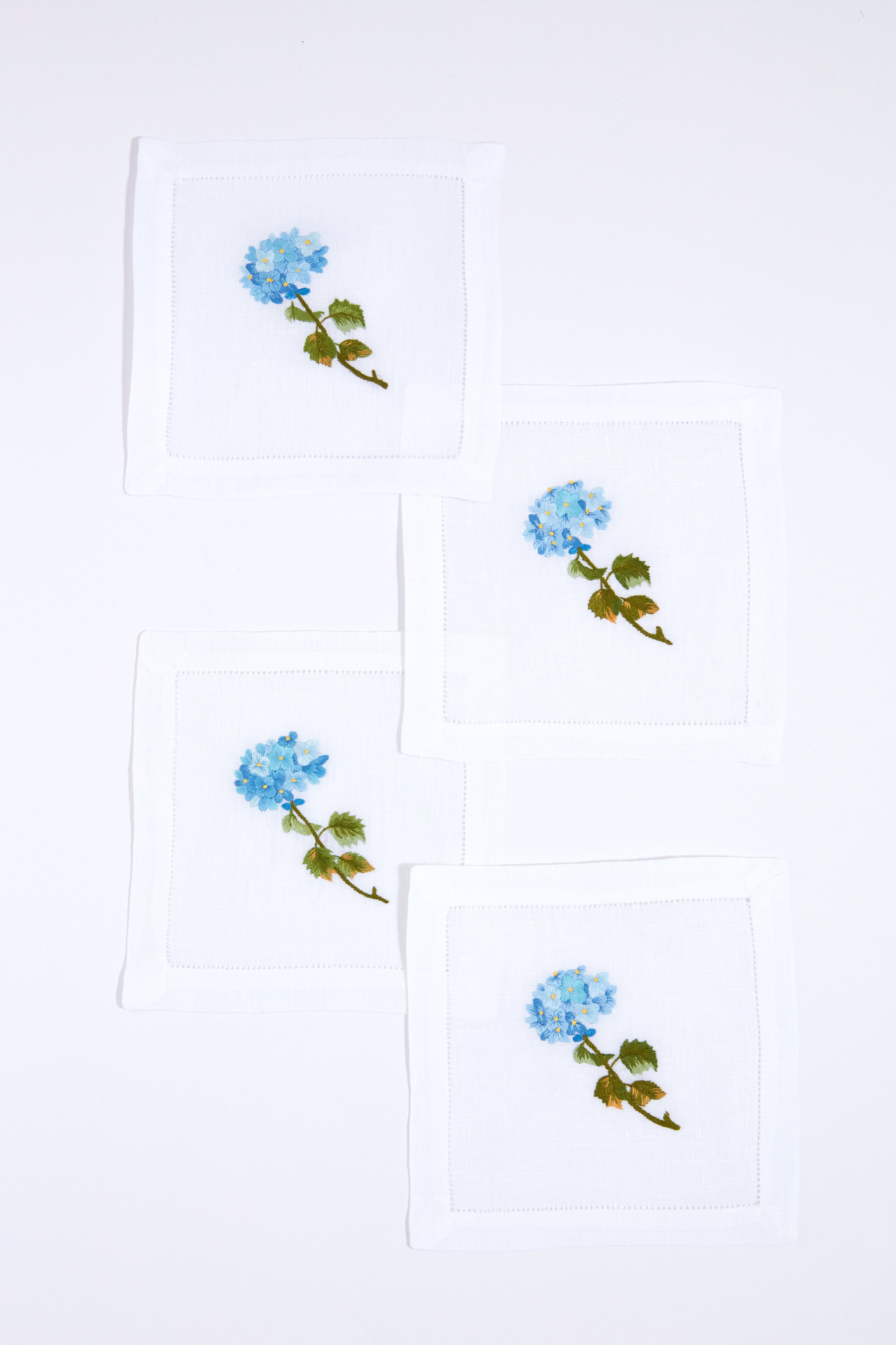 Set of four white linen napkins by Tuckernuck Home, each hand-embroidered with a blue flower and green leaves. Ideal as hydrangea-themed cocktail napkins or a thoughtful hostess gift.