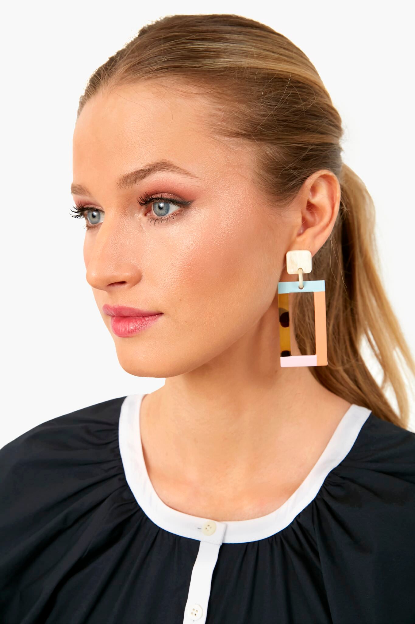 A woman with light brown hair in a ponytail wears a black top with a white collar and Sunshine Tienda's large, geometric, hand-painted horn earrings that are cruelty-free and lightweight, looking off to the side.