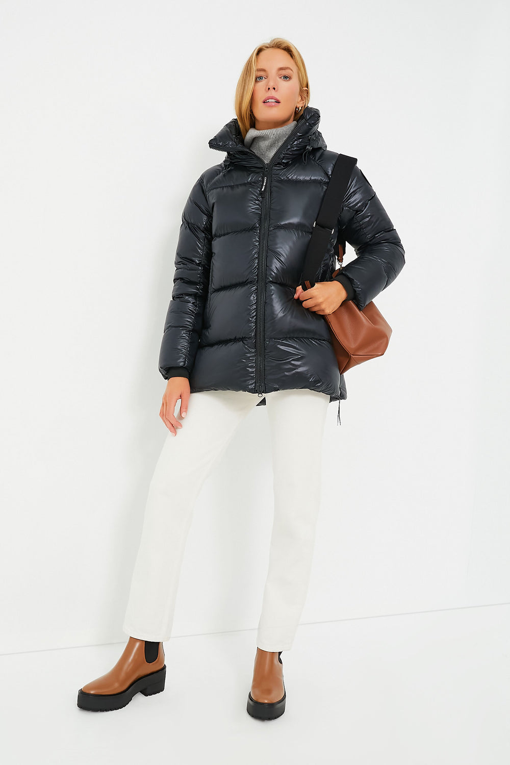 Wearing a glossy black Canada Goose jacket, white pants, and brown ankle boots, this person carries a brown shoulder bag.