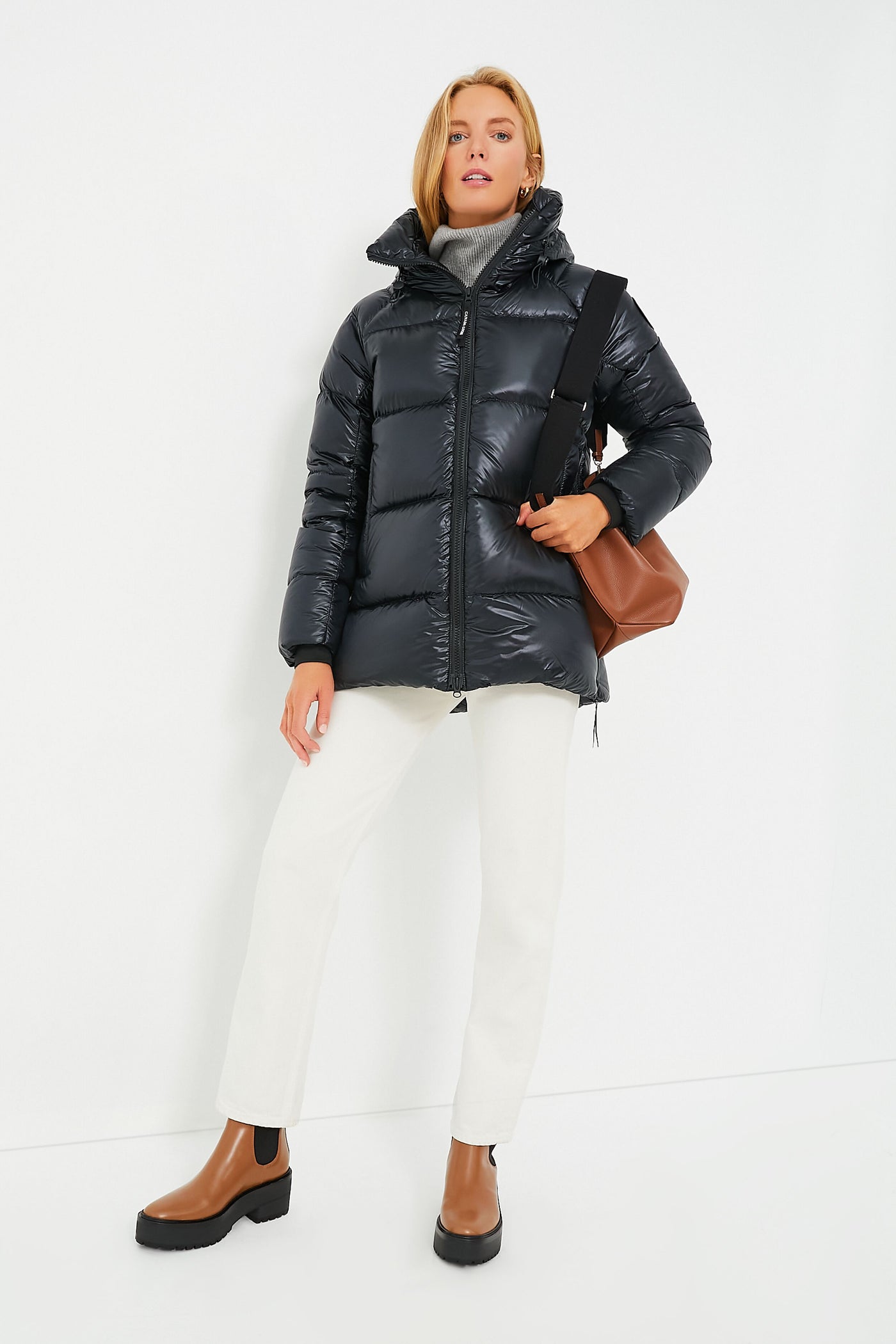 Wearing a glossy black Canada Goose jacket, white pants, and brown ankle boots, this person carries a brown shoulder bag.