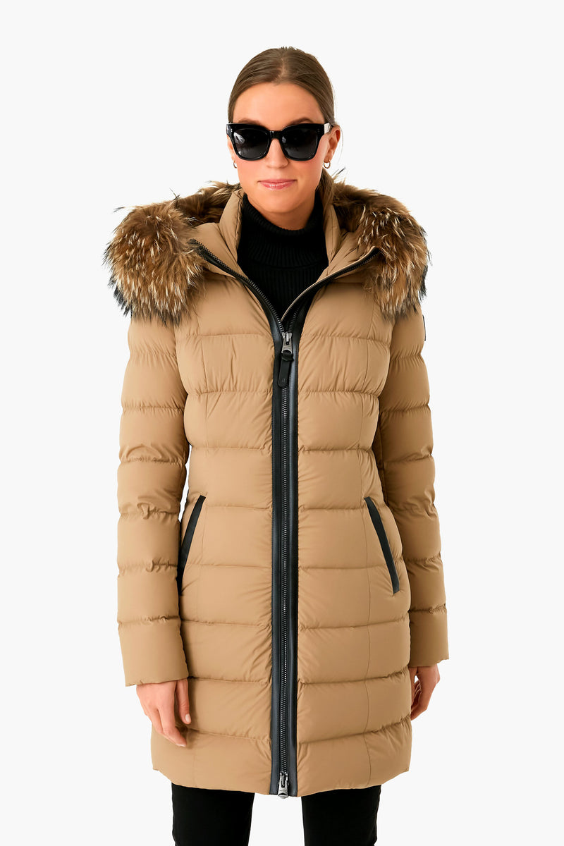 A woman wears a tan MACKAGE parka with a fur-trimmed hood, paired with a black turtleneck, black pants, and black sunglasses.