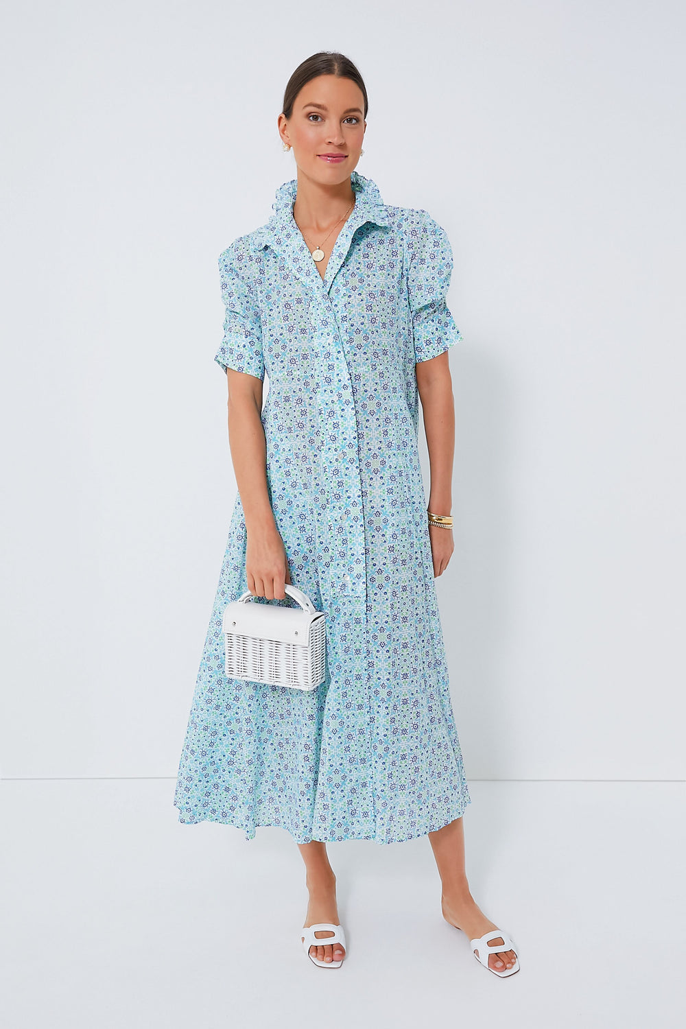A woman wears a light blue floral Thierry Colson Venetia Dress styled as a maxi, paired with white sandals and a white woven handbag.