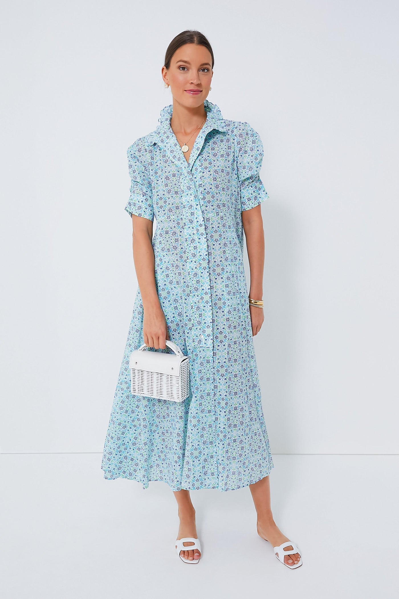 A woman wears a light blue floral Thierry Colson Venetia Dress styled as a maxi, paired with white sandals and a white woven handbag.