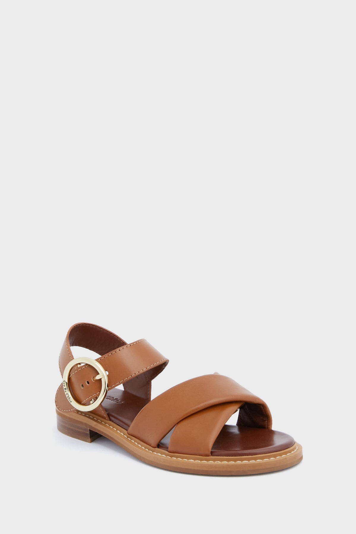 See by Chloe brown sandals with a low heel, wide crisscross toe straps, an ankle strap, and a large round gold buckle on the side.