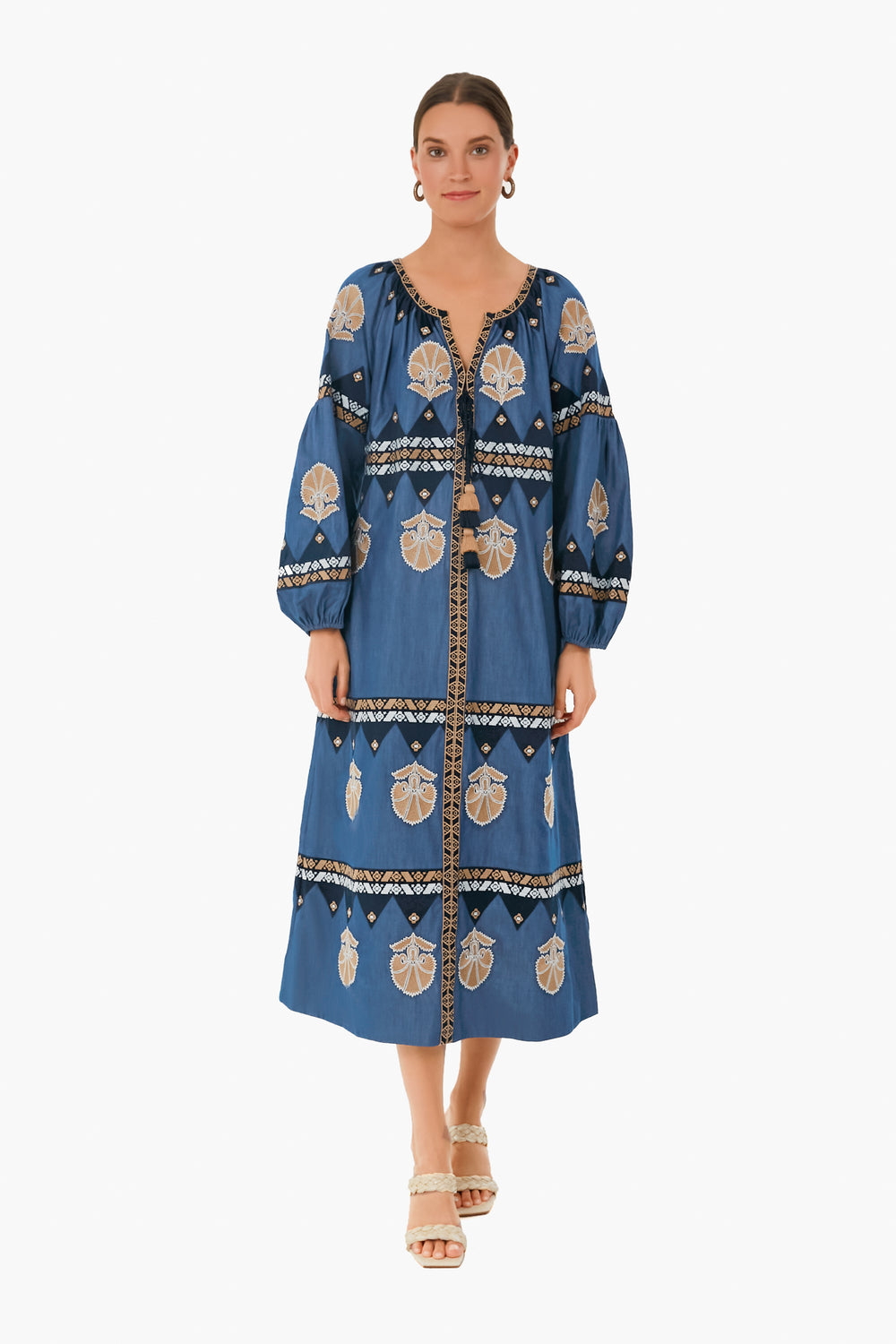 A woman faces forward in a chambray blue Figue long-sleeve embroidered midi dress with beige and black patterns, paired with beige heeled sandals.