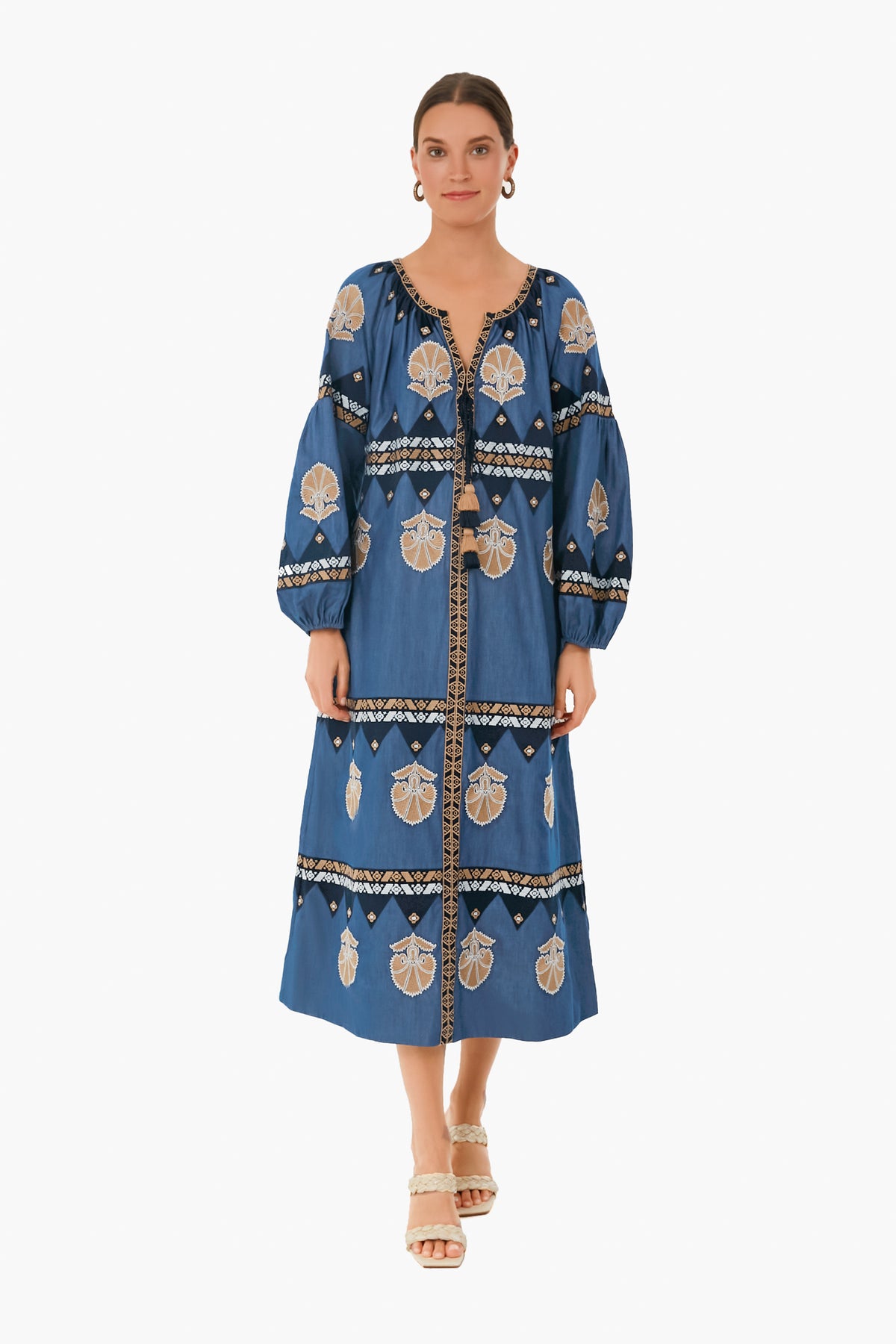 A woman faces forward in a chambray blue Figue long-sleeve embroidered midi dress with beige and black patterns, paired with beige heeled sandals.
